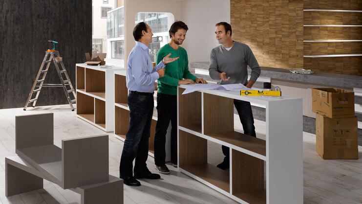 Planungsgespräch für Messe-/Showroomaufbau zwischen drei Männern mit unfertigen Mobiliar im Hintergrund